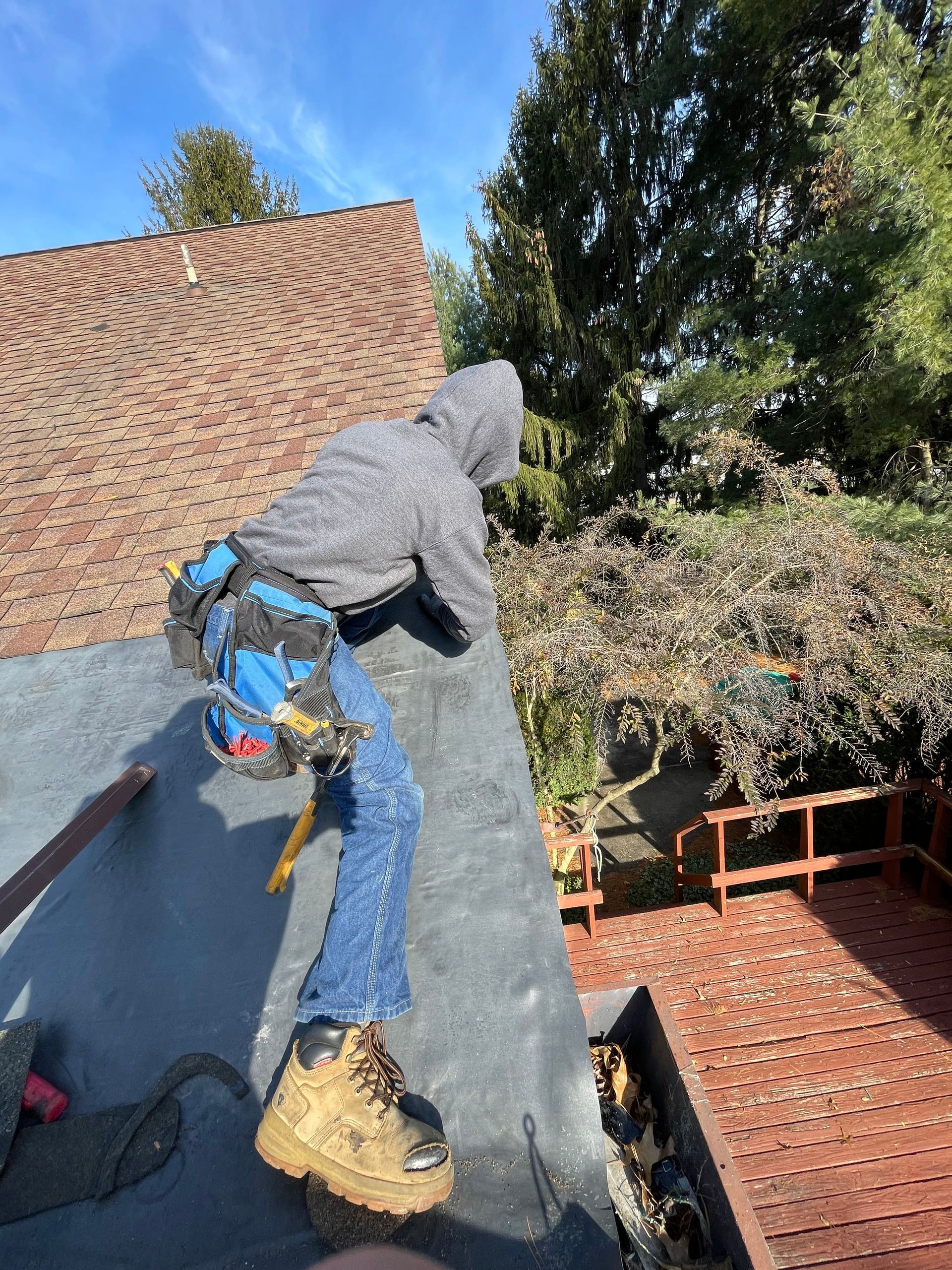 Person in gray hoodie on a roof, near a deck and trees, wearing a tool belt and boots.