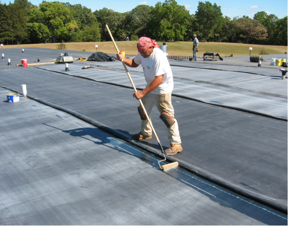 Person rolling a seam on a flat, black roofing membrane with a long-handled roller on a rooftop.