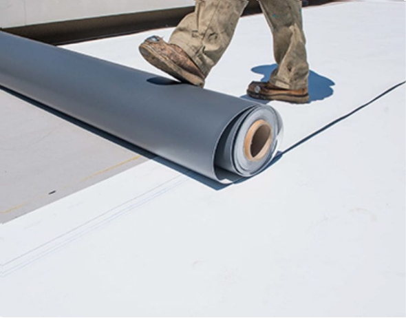 Person unrolling a roll of gray roofing material on a white rooftop, wearing work boots and tan pants.