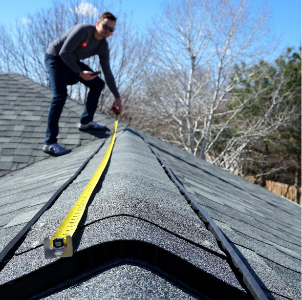 Man measures a roof ridge with a yellow tape measure under a blue sky.