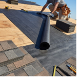 Roofer installing black roofing underlayment on wooden roof. Shingles and tools are also present.
