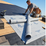 Roofer installing roofing underlayment on a wooden roof deck under a bright sky.