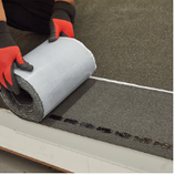 Person wearing red gloves unrolling self-adhesive roofing material along a white edge.