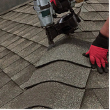 Roofer using a nail gun to install gray shingles on a roof. Red gloves visible.