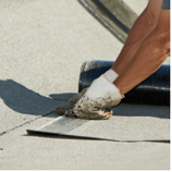 Person installing roofing material; hands wearing gloves laying black roll on gray surface.