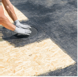 Person wearing gloves installing black roofing material around a circular drain on light-colored wooden sheeting.