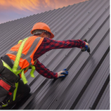 Roofer wearing safety gear, working on a gray corrugated metal roof, holding a drill, against a sunset sky.