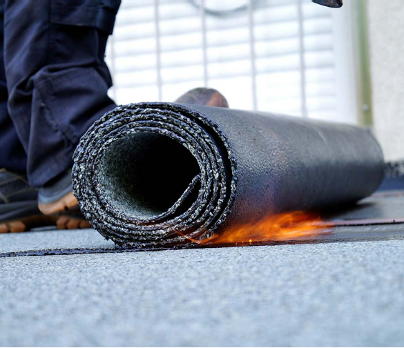 Roofer using a torch to install rolled roofing material on a gray surface. Flames visible.