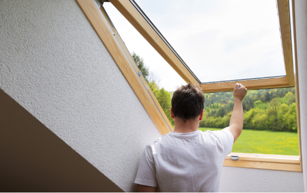Man opening a wooden skylight, overlooking a green landscape on a sunny day.