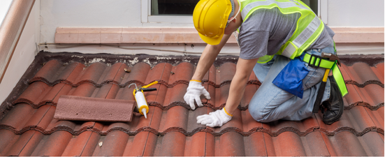 A worker on a red tile roof, wearing a hard hat, and safety vest, repairing with a caulking gun.
