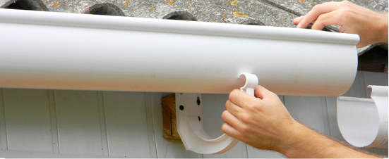 Person installing a white gutter with a downspout, on a white wall.