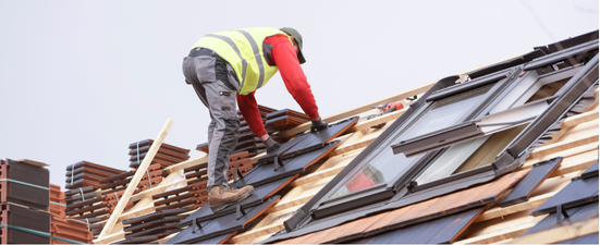 Roofer installing roof tiles on a house. He is wearing a yellow vest and red long sleeve.