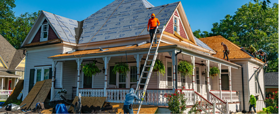 Workers repairing a house roof, one on a ladder, another on the roof.  The house is white with a porch.