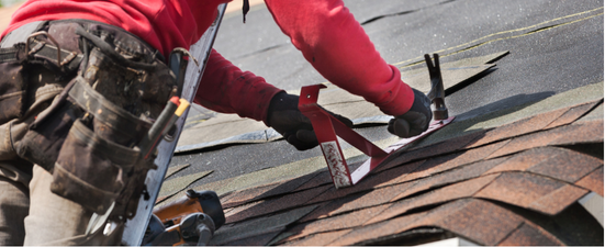Roofer in red shirt working on roof, using a hammer and tool.