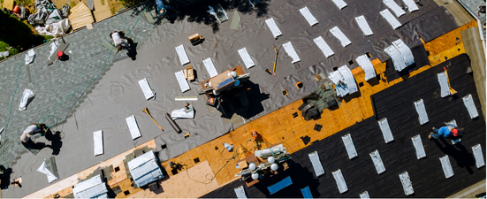 Roofers working on a partially-completed roof, with white paper strips and tools scattered on the surface.