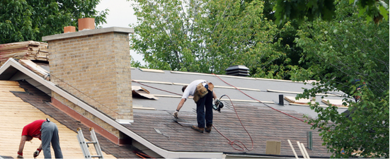 Roofers working on a roof, near a chimney and trees. One roofer is bent over with his back to the camera.