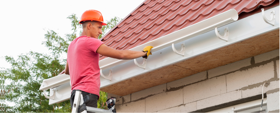 A person wearing a hard hat, cleaning a white gutter on a brick building with a red roof.