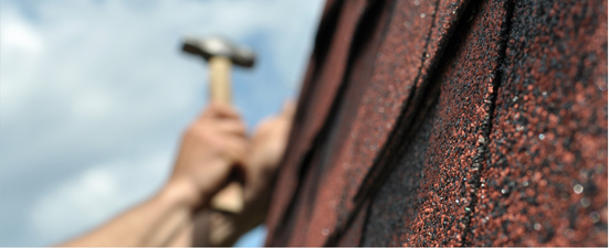 Person hammering roofing shingles against a cloudy sky.