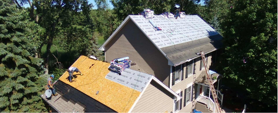 A house being worked on with a partially replaced roof. Green trees surround the house.
