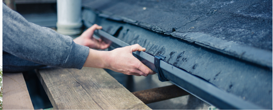 A person wearing a hard hat, cleaning a white gutter on a brick building with a red roof.