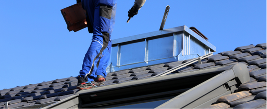 A person in blue overalls on a roof, near a skylight, holding a clipboard and tool, under a blue sky.