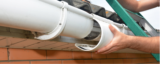 A person installing a white gutter on a brick house, next to a green ladder.