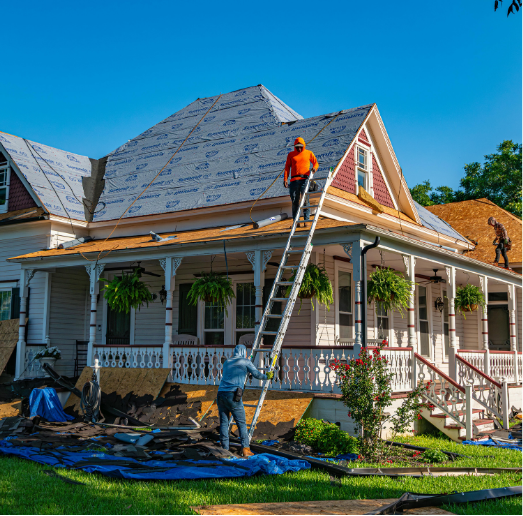 Roofers replacing shingles on two-story house with porch, one on roof and one on ladder, blue tarp on ground.