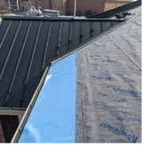 Roof under construction, showing wooden planks laid over a dark underlayment, with trees in the background.