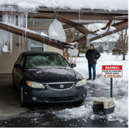 Snow-covered car under a collapsed carport. Man stands beside a warning sign.