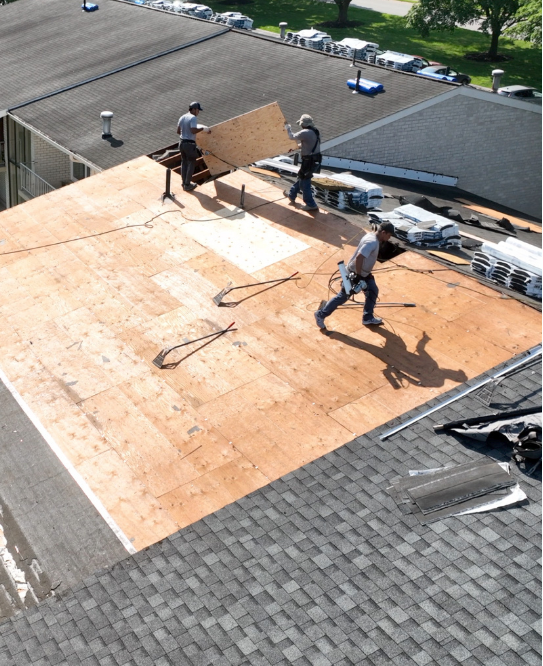 Three roofers working on a flat roof, installing plywood. Gray and tan materials. Sunny day.