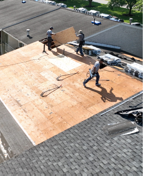 Roofers installing plywood on a flat roof. They are wearing harnesses, laying a large board near a hole.