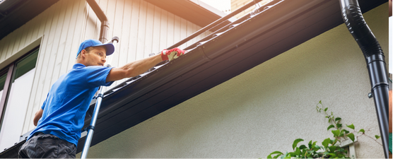 Person installing a white gutter on a brick building, holding a downspout piece.