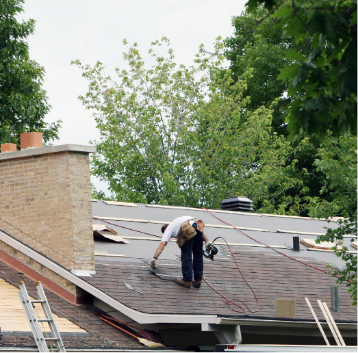 Roofer working on a roof, near a brick chimney and trees in the background, cloudy sky.
