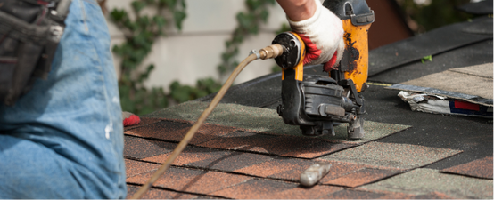 Person using a nail gun on a roof, with orange and gray shingles.