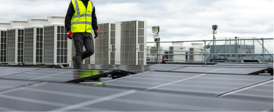Person in a safety vest on a rooftop with solar panels, air conditioning units, and a cloudy sky.