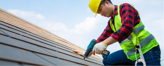 Roofer in safety gear, drilling into a tiled roof, against a blue sky.
