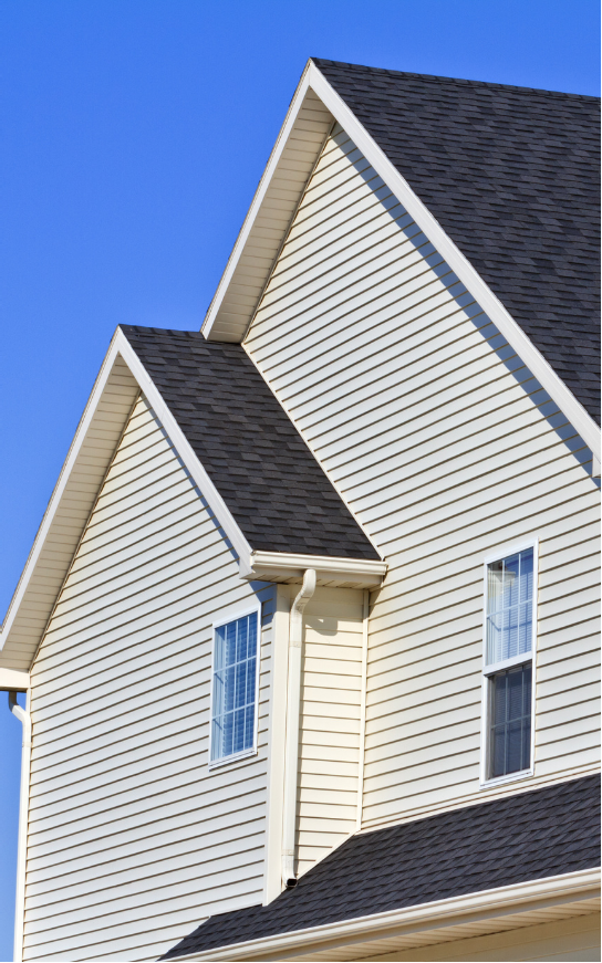 Side view of a house with light siding, dark roof, and blue sky. Two windows are visible.