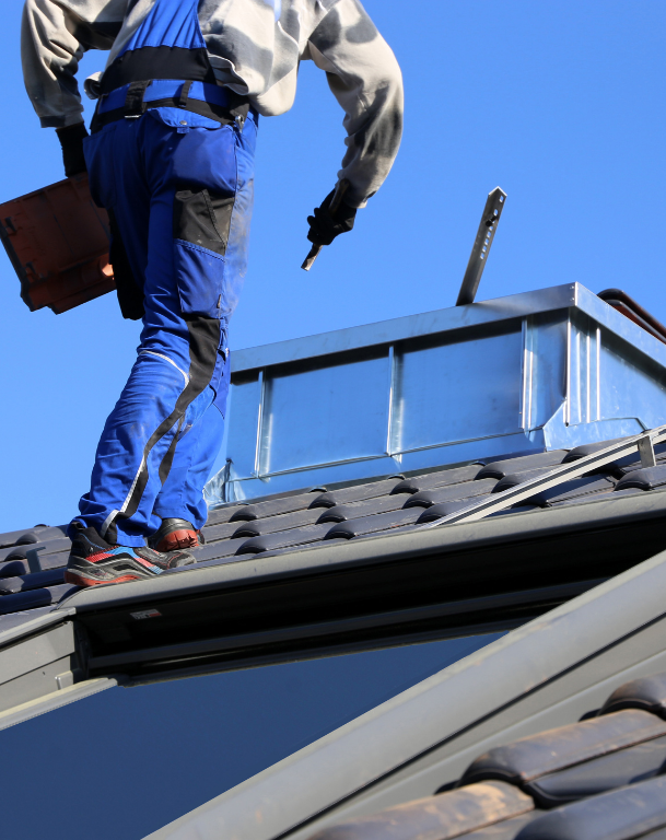 Roofer in blue overalls on a tiled roof by a metal vent, holding a tile and a tool, working on a bright, sunny day.