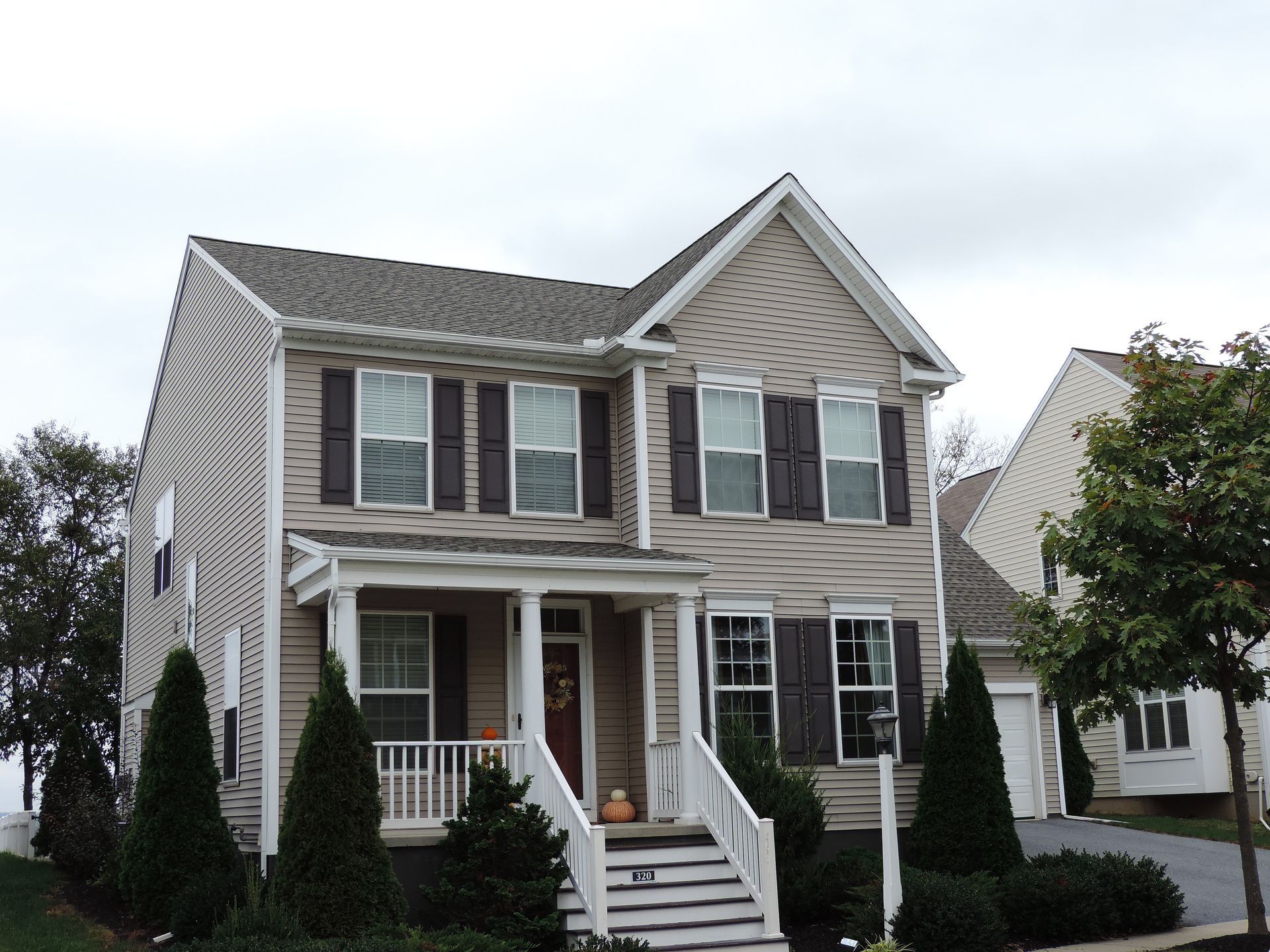 Two-story house with tan siding, brown shutters, white porch, and a gray roof on an overcast day.