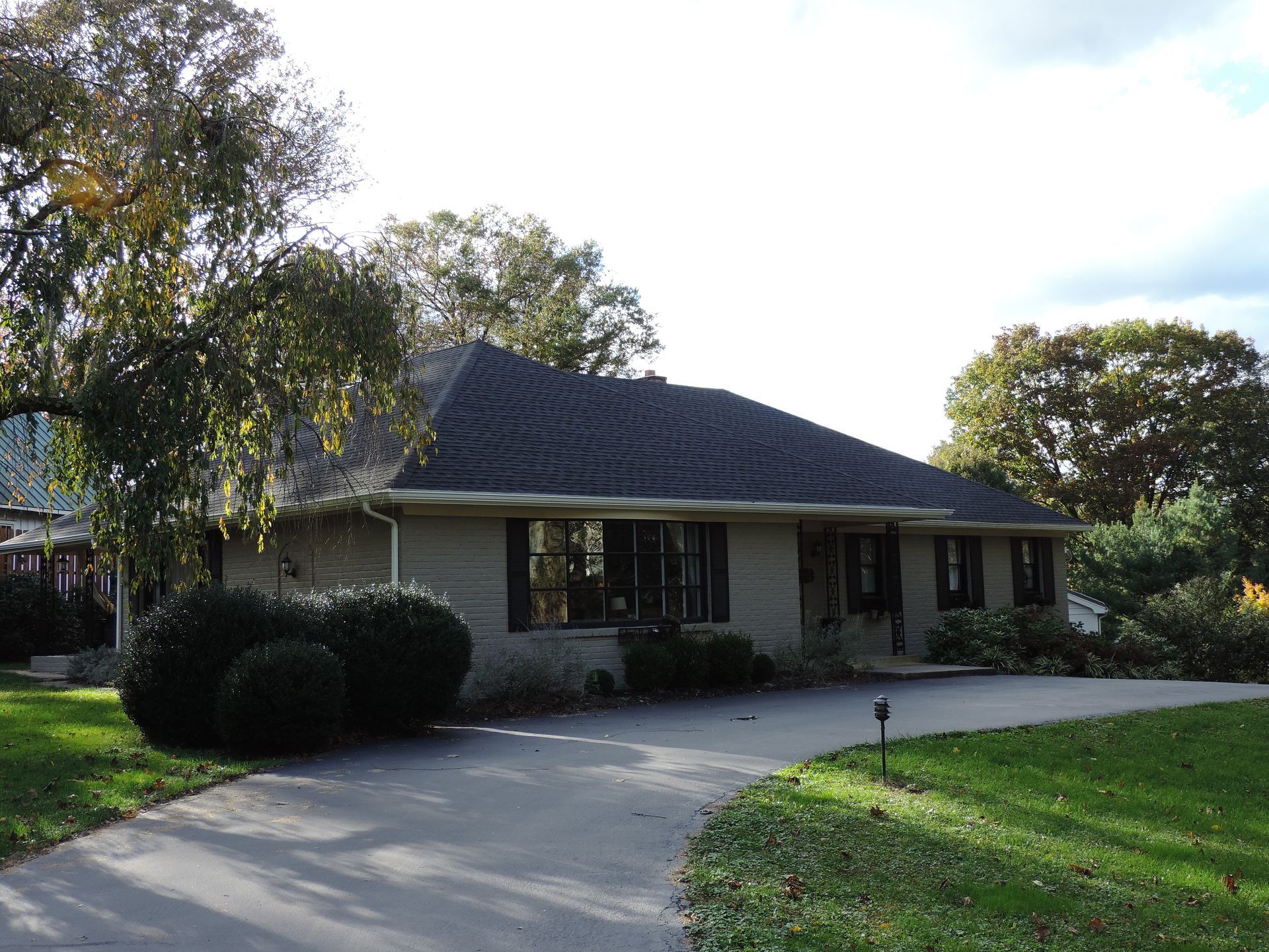 A single-story beige house with a dark roof and a curved driveway. Trees and green grass surround it.