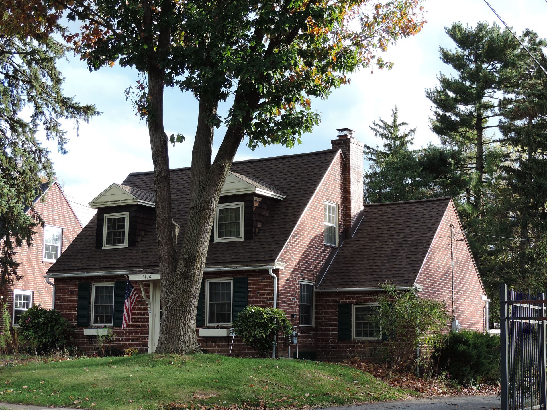 Red brick house with dark roof, dormers, and chimney. Tree in front, green lawn, blue sky.