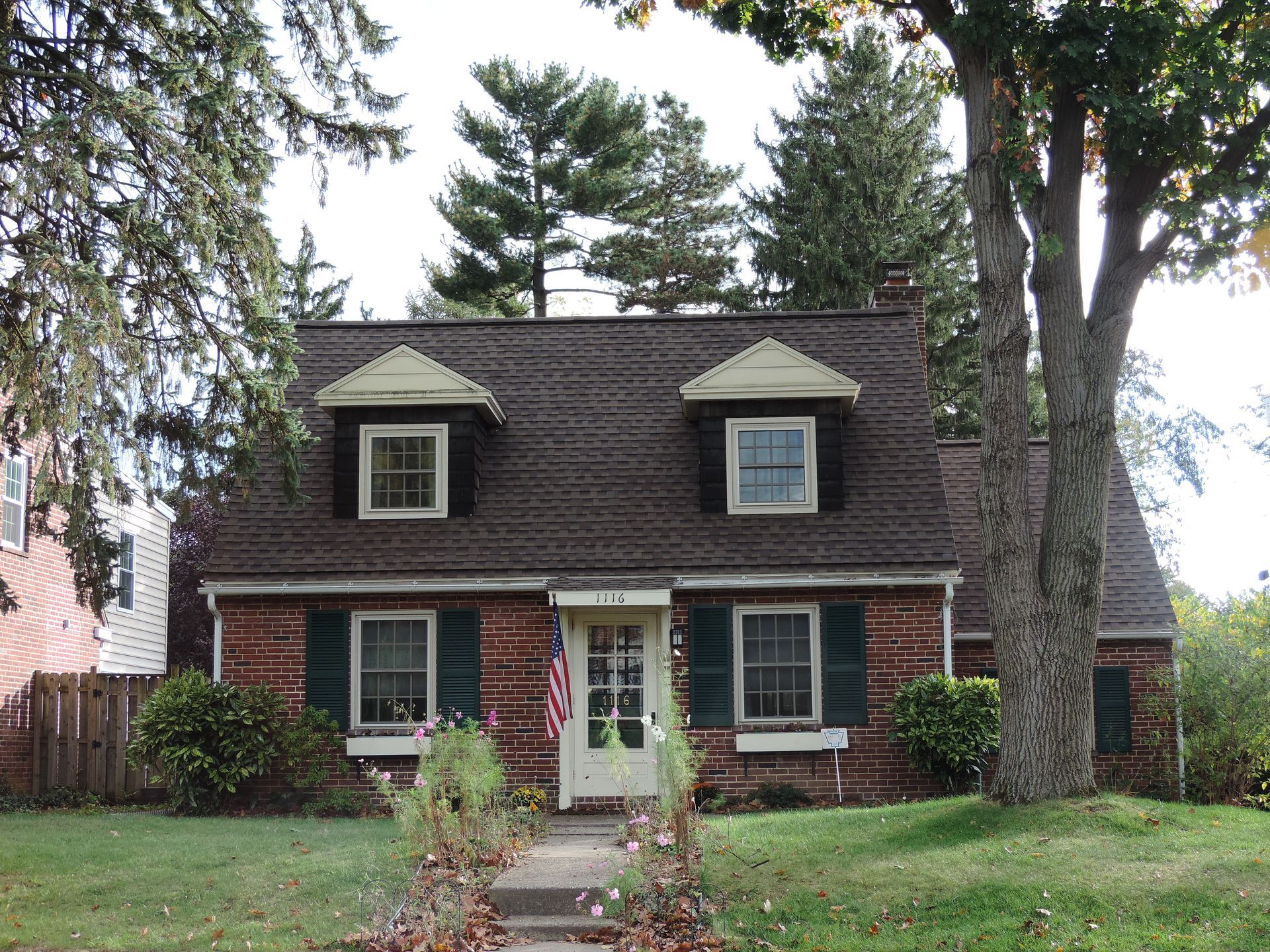 Brick house with two dormers, green shutters, American flag, and a tree on the right.