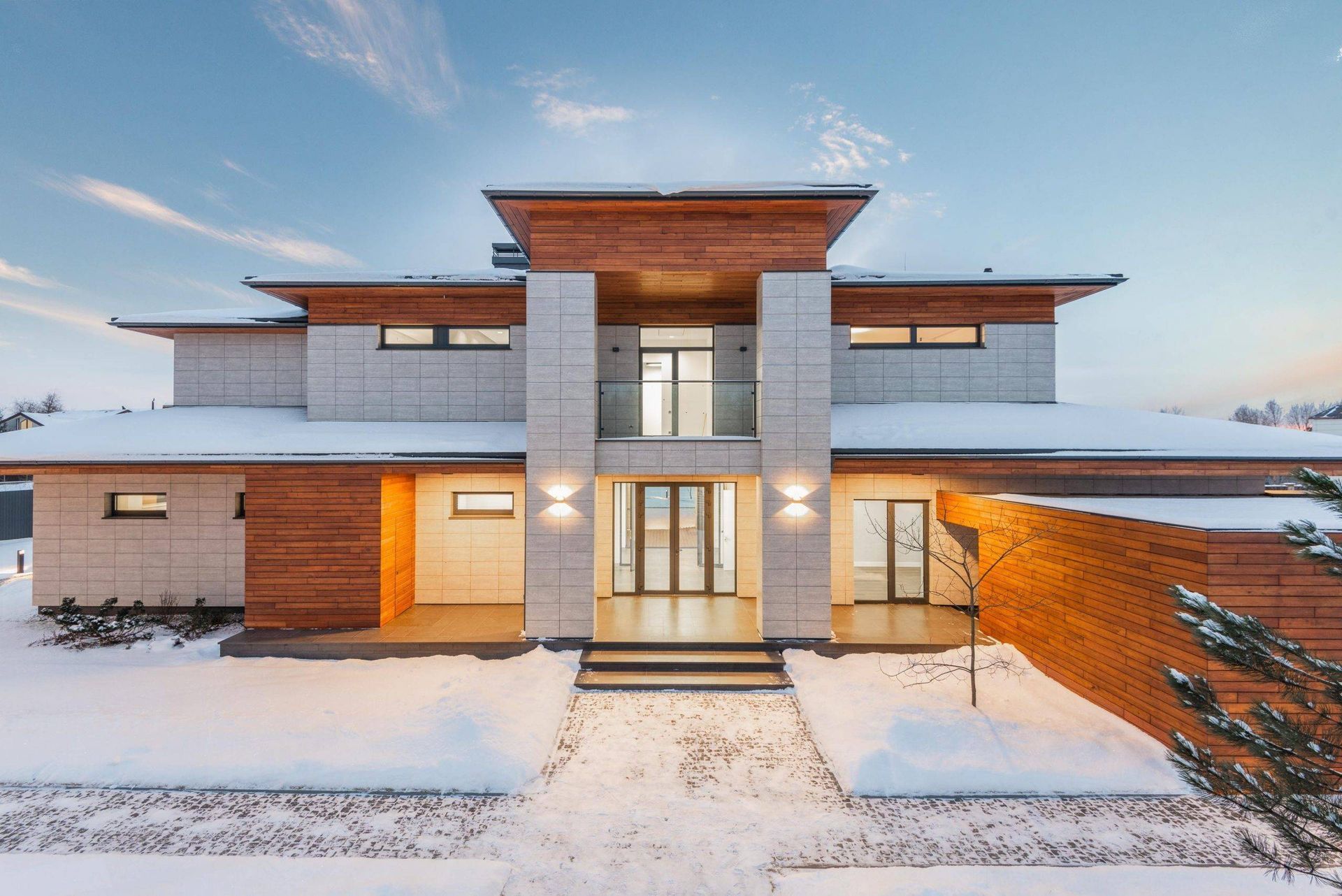 Modern two-story house with light stone and wood accents, snow on the roof and ground, at dusk.
