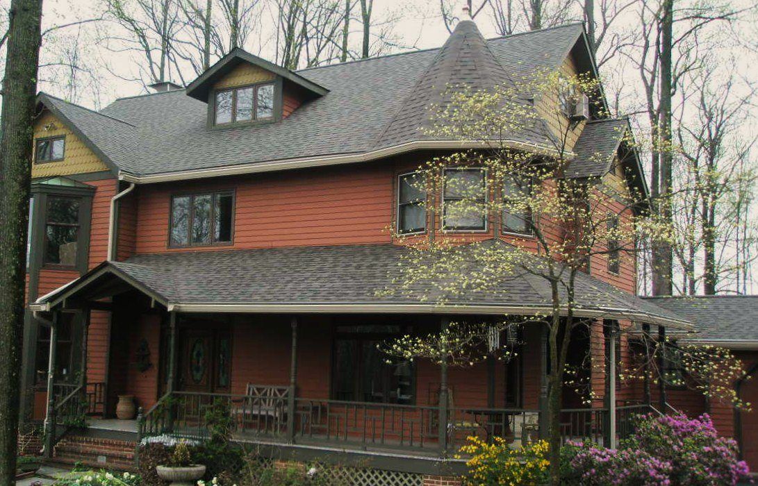 Two-story orange house with a wraparound porch, set among trees. Features include a dormer window and dark gray roof.