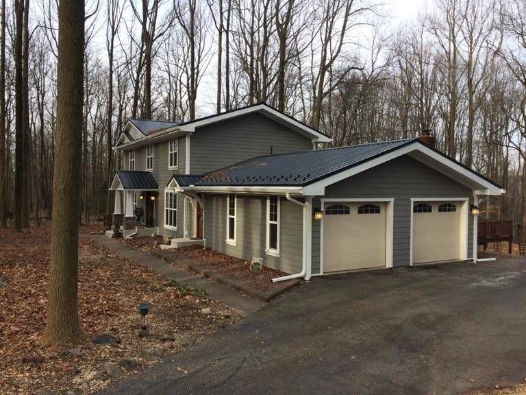 Gray two-story house with black roof and two-car garage set in a wooded area; driveway in front.