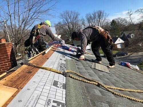Two roofers working on a rooftop, securing shingles. One wears a safety harness. Blue sky in the background.
