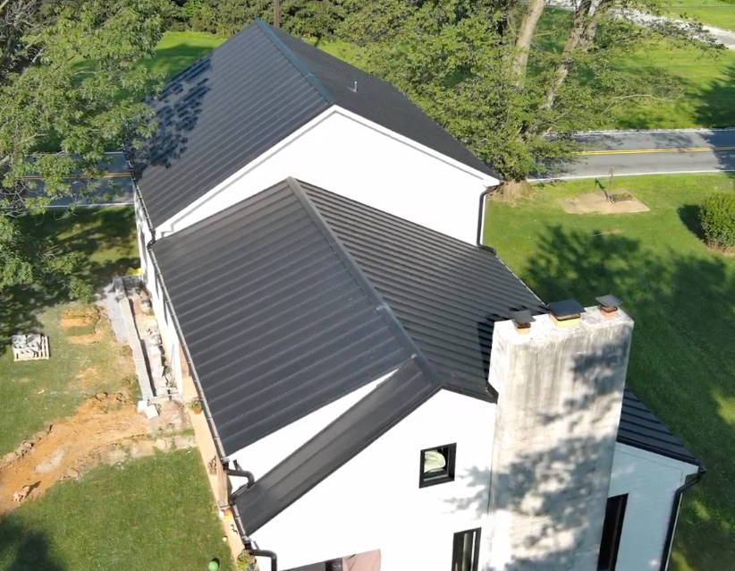 Black metal roof on a white house, aerial view. Exterior features a chimney, green lawn, and trees.
