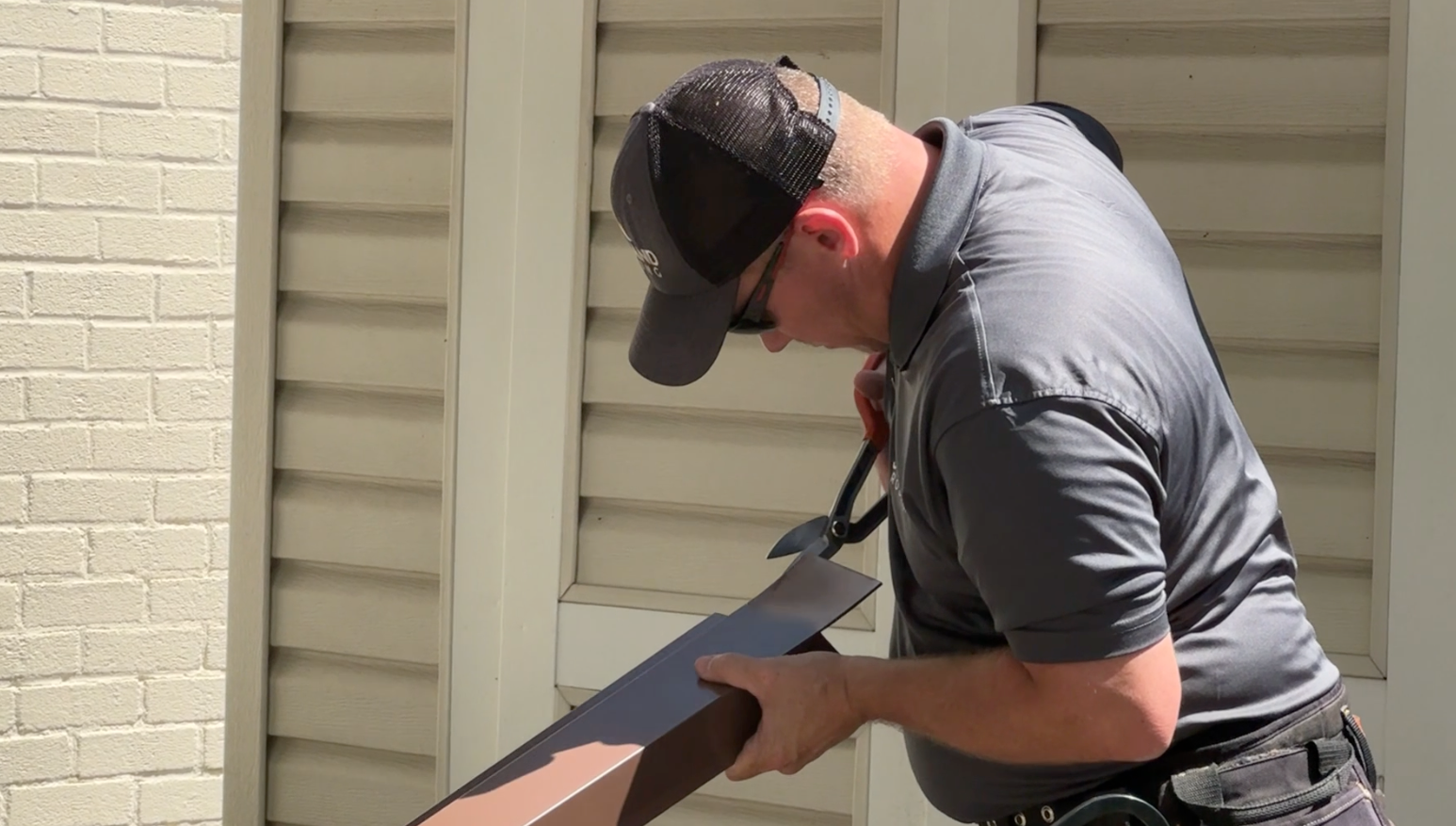 Man cutting a brown piece of material near a building with light siding. He wears a hat and sunglasses.