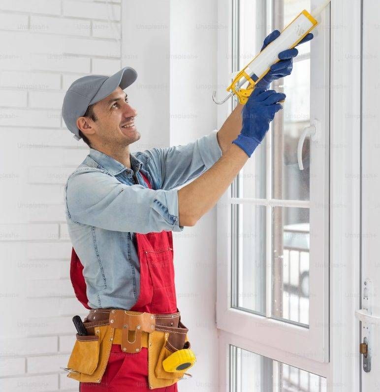 Construction worker caulking a white window frame with a sealant gun, wearing a cap, gloves, and tool belt.