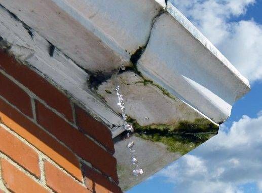 Leaking white gutter on a brick building with water dripping, showing algae growth.